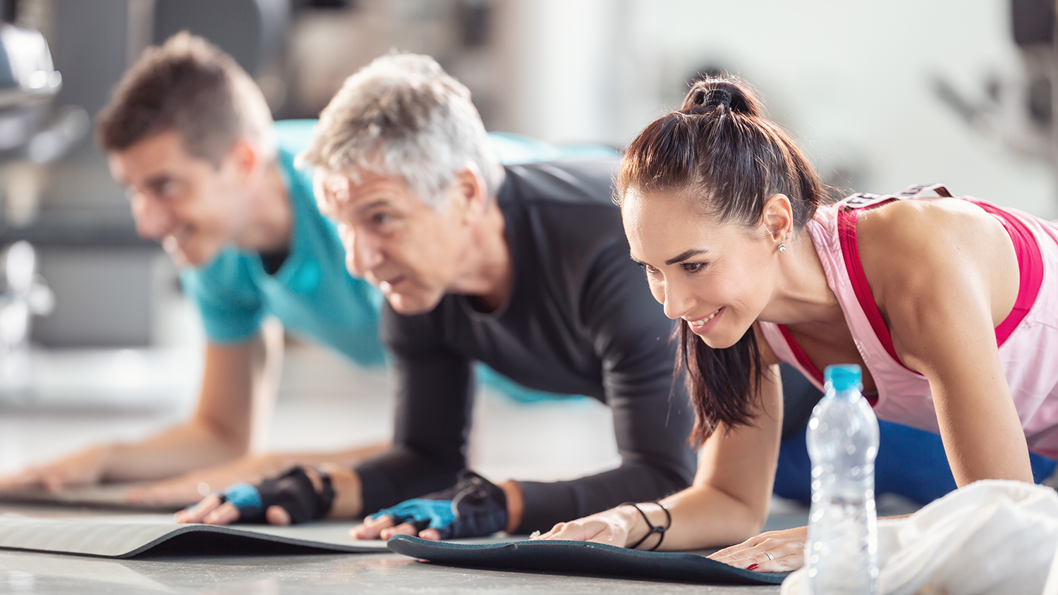 Two men and a woman doing elbow planks on mats inside a gym with a plastic water bottle in the front.