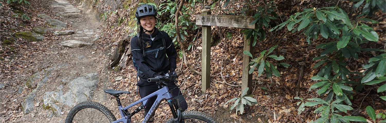 A young woman stading outside wearing a bike helmet and besides a bike.