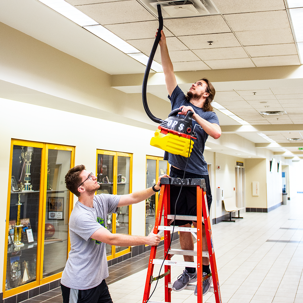 A guy cleans the AC vents while another holds the ladder.